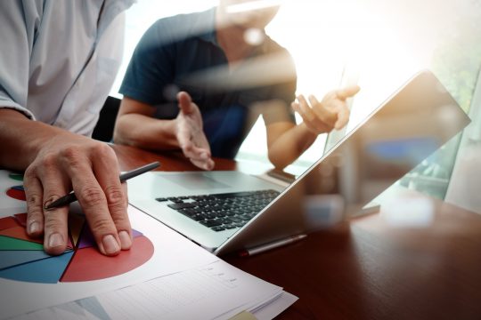 business documents on office table with smart phone and laptop computer and graph business with social network diagram and two colleagues discussing data in the background